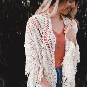 A woman wearing a white shawl decorated with the intricate crochet pineapple stitch, with fringed edges. She stands in an outdoor green space, surrounded by greenery and sunshine.