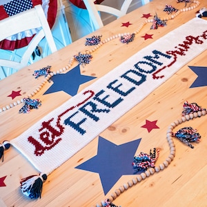 May include: A white crocheted table runner with the words "Let Freedom Ring" in blue and red yarn. The runner is on a wooden table with a blue and white star garland and red stars.