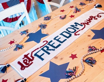 May include: A white crocheted table runner with the words "Let Freedom Ring" in blue and red yarn. The runner is on a wooden table with a blue and white star garland and red stars.