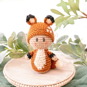 May include: A crocheted fox doll wearing a brown and white hat with black ears. The doll is sitting on a wooden base with a beaded edge.