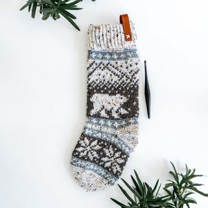 May include: A white and brown knitted stocking with a polar bear pattern. The stocking has a brown leather loop at the top and a white X stitched on the leather.