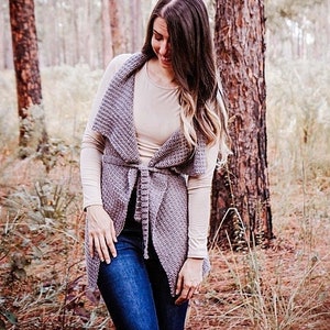 May include: A woman wearing a grey knitted vest with a tie belt, a beige long-sleeved shirt, and blue jeans. She is standing in a woodland with tall trees.