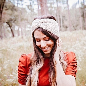 Peut inclure: Une femme aux longs cheveux blonds porte un bandeau en crochet blanc. Elle sourit et regarde vers le bas. Elle porte une chemise rouge et se tient dans un champ d'herbe haute.