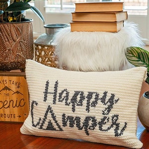 May include: A white crocheted pillow with the text "Happy Camper" in black embroidery. The pillow is sitting on a white faux fur ottoman with a stack of books on top. There is a wooden sign with the text "Always Take The Scenic Route" in front of the ottoman.