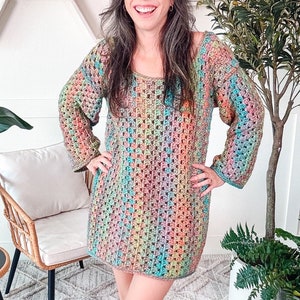 A person wearing a colorful granny crochet dress and unique jewellery stands indoors near a chair and various plants, smiling at the camera.