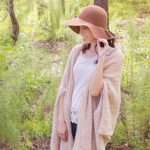 A woman wearing a cream crochet shawl wrap and hat, in a field of greenery.