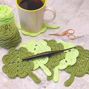 A crochet project in progress with green yarn, showcasing several completed leaf-shaped pieces intended for a coaster, alongside a crochet hook, scissors, and a mug on a marble surface.