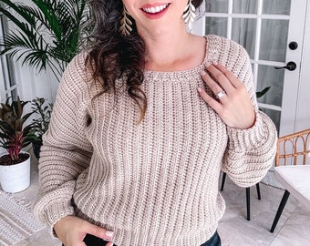 May include: A beige long-sleeved jumper featuring a vertical ribbed knit pattern, a rounded neckline, and fitted cuffs. The garment is paired with dark-coloured denim jeans and gold-toned leaf-shaped drop earrings.