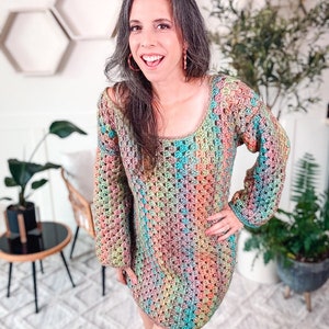 A woman stands indoors wearing a colorful, crocheted dress and unique jewelry. She is smiling, with one hand on her hip. The background features plants and hexagonal wall shelves.