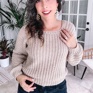 Woman in a beige crochet sweater, featuring unique jewellery earrings, posing with hand on hip.