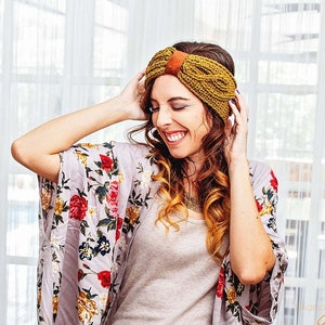 May include: Mustard yellow knitted headband with a brown leather accent. The headband is worn by a person wearing a floral kimono with red, yellow, and white flowers. The background is a window with white curtains.