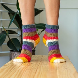 Puede incluir: Un par de calcetines a rayas de colores con un patrón morado, marrón, rojo, blanco y amarillo. Los calcetines están hechos de un tejido suave de punto y se muestran sobre una superficie de madera.