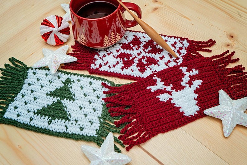 May include: Three crocheted coasters with fringe, featuring a Christmas tree, a snowflake, and a reindeer. The coasters are in green, white, and red.
