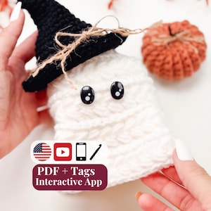 May include: A white crocheted ghost figure wearing a black crocheted witch hat with a jute twine bow. The ghost has two black plastic safety eyes. A small orange crocheted pumpkin sits nearby. Text on the image reads: PDF + Tags Interactive App.