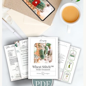 May include: A digital PDF pattern for a Wheat Stitch Pickle Ornament by Briana K Designs, featuring printed pages with crochet instructions, abbreviations, supply lists, and gift tags, displayed alongside a tablet showing a video tutorial.