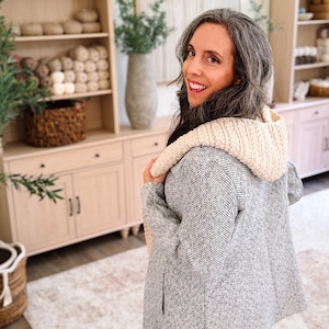 May include: A gray and white herringbone patterned jacket with a cream-colored, thick, ribbed knit hood. The jacket features long sleeves and a straight hemline, providing a textured appearance with a contrasting soft, chunky knit hood detail.