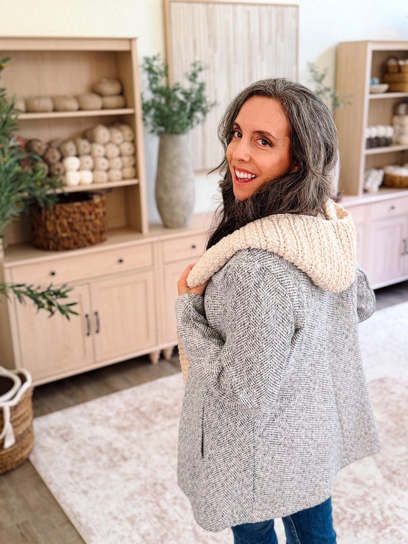 May include: A gray and white herringbone patterned jacket with a cream-colored, thick, ribbed knit hood. The jacket features long sleeves and a straight hemline, providing a textured appearance with a contrasting soft, chunky knit hood detail.