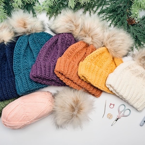 Row of crochet beanies in green, navy, teal, purple, orange, yellow and cream with pom poms and tools