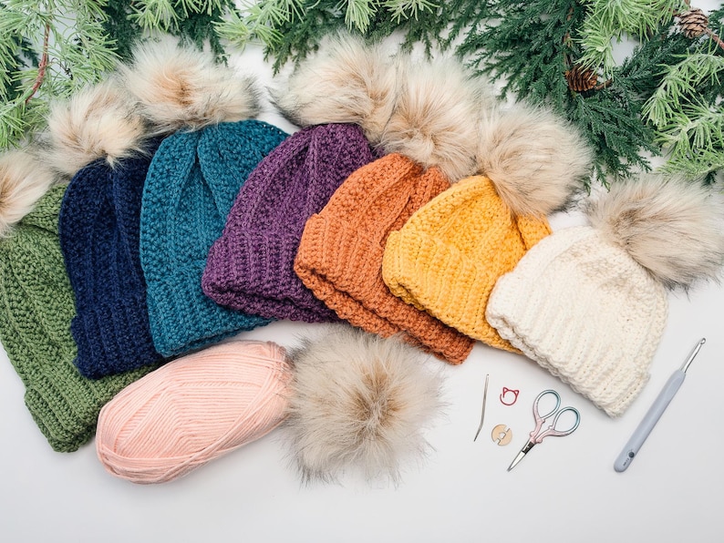 Row of crochet beanies in green, navy, teal, purple, orange, yellow and cream with pom poms and tools