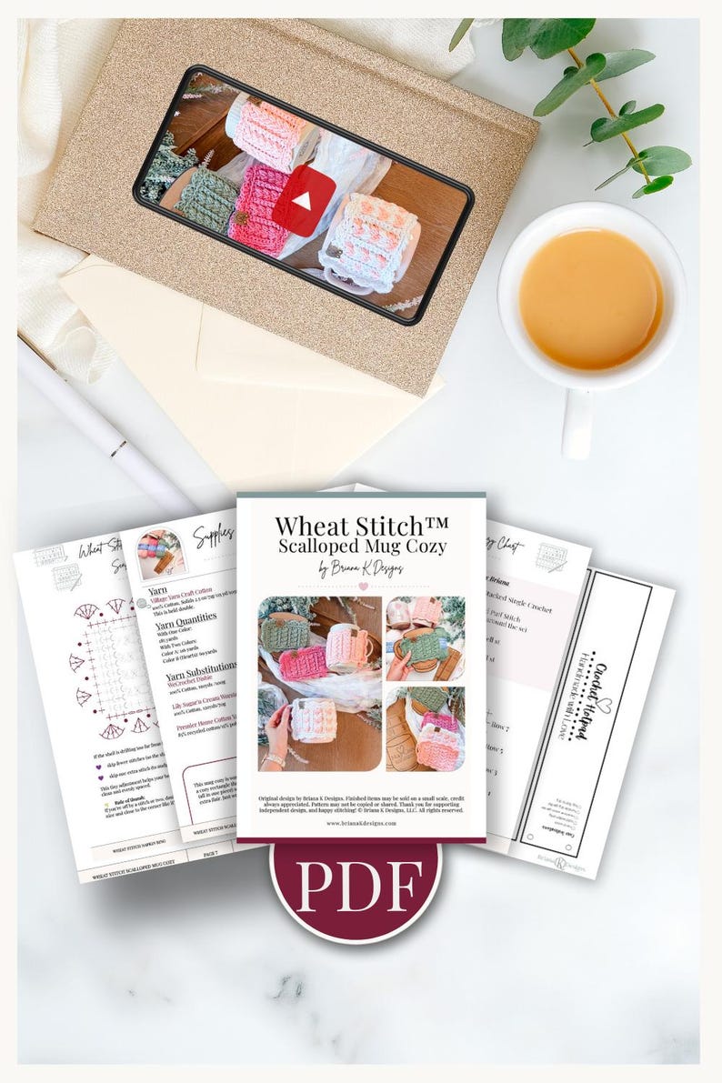 May include: An overhead shot features a collection of crochet patterns and supplies. A phone displays a video of colorful crochet mug cozies. The main pattern is titled "Wheat Stitch Scalloped Mug Cozy" and includes a PDF download. A cup of tea sits nearby.