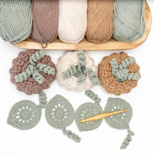 May include: Five skeins of yarn in sage green, brown, cream, and tan sit in a wooden tray above three crocheted pumpkins in matching colors. Below are four sage green crocheted leaves and a wooden crochet hook on a plain white background.