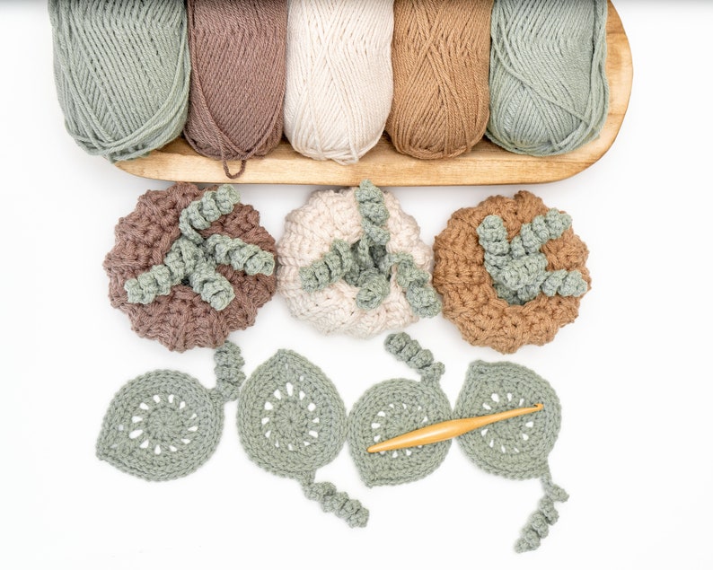 Pumpkin Coasters crochet pattern: crochet pumpkins and sage leaf coasters below five skeins of neutral yarn, with a wooden crochet hook