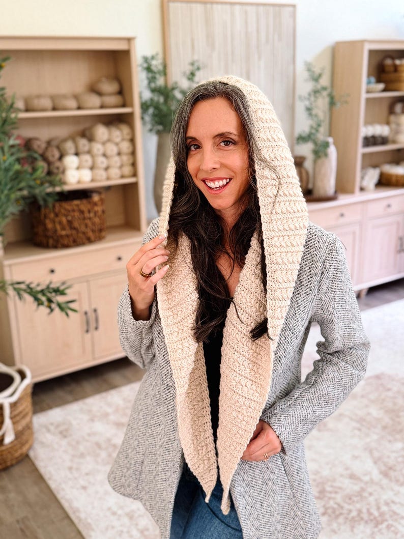 May include: A cream-colored crochet hood with a textured, ribbed stitch pattern. The hood is designed to be worn over the head and drapes down the front of the chest, featuring a wide, thick border that frames the face with a consistent knit texture.