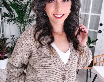 Op de afbeelding: Een beige en bruin gehaakt vest met een knoopsluiting aan de voorkant. Het vest wordt over een witte top gedragen. De mouwen zijn lang en de pasvorm is cropped. De achtergrond bevat planten en een witte deur.