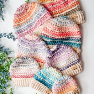 May include: A collection of seven crocheted beanies in various sizes, each featuring horizontal stripes of variegated yarn in pastel and vibrant colors. Every hat has a solid cream-colored ribbed brim and a small brown leather tag.