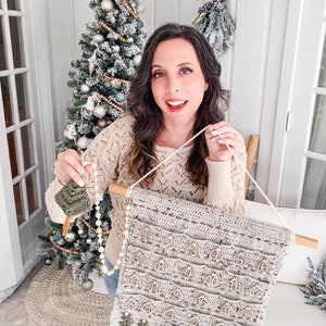 May include: A handmade crochet advent calendar in light gray with wooden ornament-shaped pockets numbered 1-25. The calendar is suspended from a wooden dowel with a white string. Small green crochet trees and a small green square are also visible.