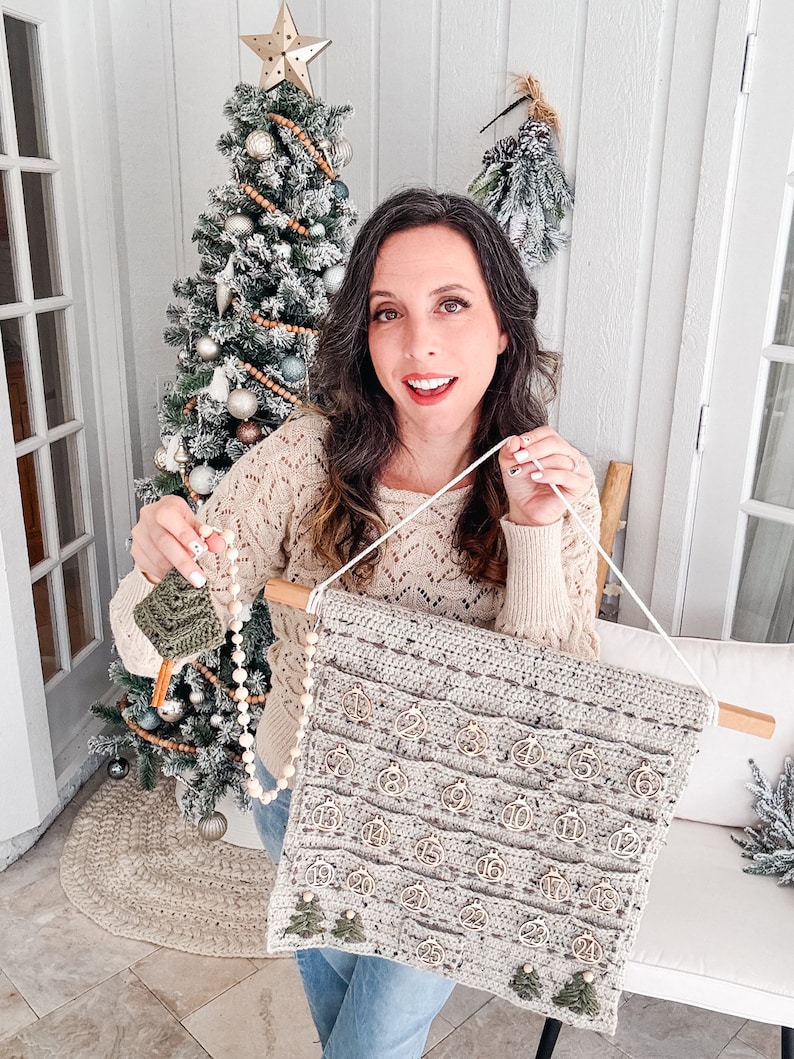 May include: A handmade crochet advent calendar in light gray with wooden ornament-shaped pockets numbered 1-25. The calendar is suspended from a wooden dowel with a white string. Small green crochet trees and a small green square are also visible.