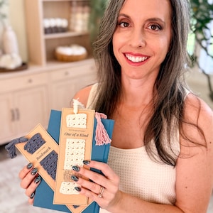 May include: Two rectangular kraft paper cards holding crocheted bookmarks. One card features a dark blue crocheted bookmark with a matching tassel and the text Bookmark. The other card holds a cream crocheted bookmark with a pink tassel and the text One of a Kind.