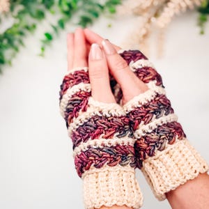May include: Hand-crocheted fingerless gloves in cream, burgundy, and purple. The wrist cuffs are a solid cream colour, while the main body features a textured pattern with stripes of cream, burgundy, and purple. The gloves are designed to keep hands warm while leaving fingers free.