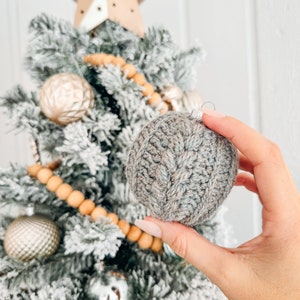 May include: A round, grey, crocheted Christmas ornament featuring a textured cable knit pattern. The ornament has a silver metal cap at the top and is held against a flocked Christmas tree decorated with gold ornaments and a star topper.