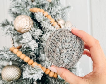 May include: A round, grey, crocheted Christmas ornament featuring a textured cable knit pattern. The ornament has a silver metal cap at the top and is held against a flocked Christmas tree decorated with gold ornaments and a star topper.