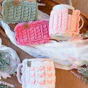 May include: Four handmade crochet mug cozies in shades of green, pink, and white. Each cozy features a textured cable knit design and a small leather tag. The cozies are displayed on a wooden tray with a white cloth and floral accents.