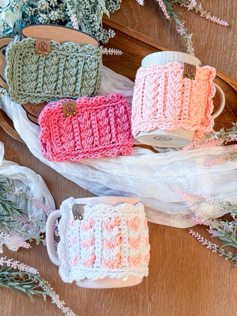 May include: Four handmade crochet mug cozies in shades of green, pink, and white. Each cozy features a textured cable knit design and a small leather tag. The cozies are displayed on a wooden tray with a white cloth and floral accents.