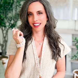 May include: A woman wearing a cream-colored, short-sleeved top and a matching bracelet. The top has a textured knit pattern. A lanyard with a colorful floral design hangs around her neck. She is smiling and wearing gold earrings.