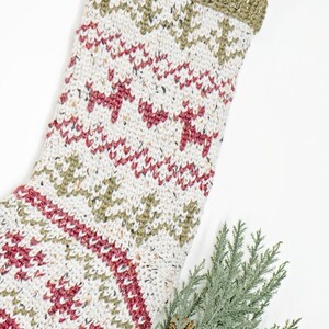 Op de afbeelding: Een gebreide kerstsok met een cr&egrave;mekleurige basis en gespikkeld garen, versierd met een herhalend patroon van rode rendieren en olijfgroene dennenbomen. De bovenkant is geribbeld olijfgroen breiwerk met een bruine leren ophanglus.