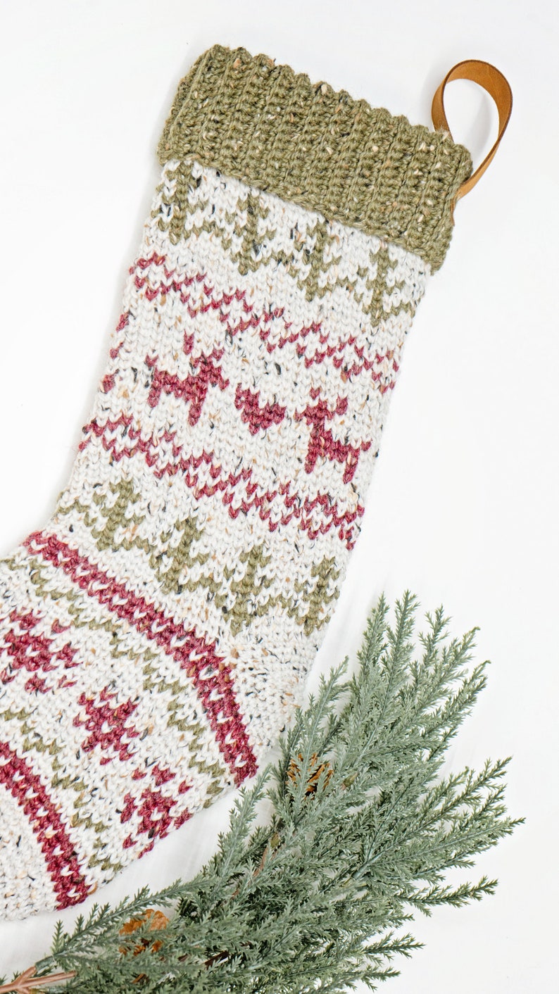Op de afbeelding: Een gebreide kerstsok met een cr&egrave;mekleurige basis en gespikkeld garen, versierd met een herhalend patroon van rode rendieren en olijfgroene dennenbomen. De bovenkant is geribbeld olijfgroen breiwerk met een bruine leren ophanglus.