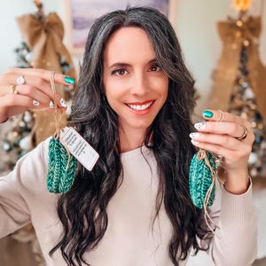 May include: Two small, textured, emerald green crocheted ornaments with brown twine loops. One ornament features a small, rectangular white paper tag with printed black text attached to the twine. The ornaments have a thick, ribbed yarn construction.