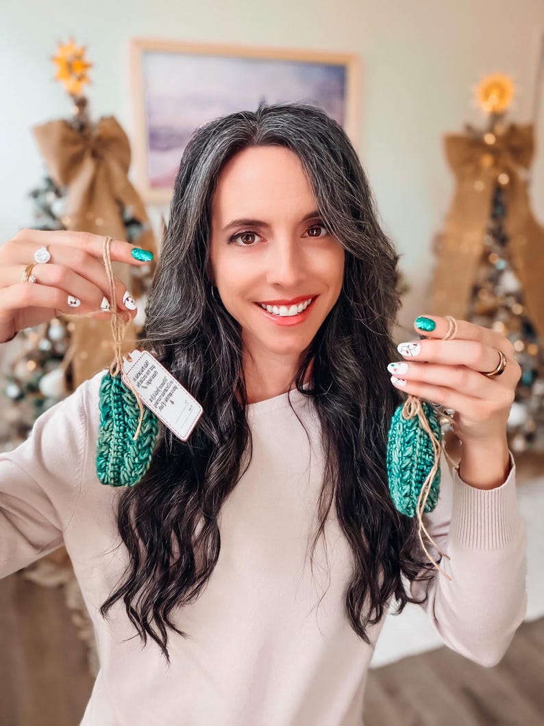May include: Two small, textured, emerald green crocheted ornaments with brown twine loops. One ornament features a small, rectangular white paper tag with printed black text attached to the twine. The ornaments have a thick, ribbed yarn construction.