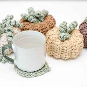 White mug on a sage crochet leaf coaster in front of four crochet pumpkins in neutral tones