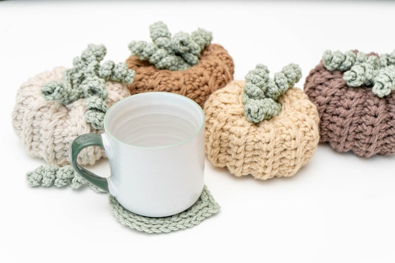 White mug on a sage crochet leaf coaster in front of four crochet pumpkins in neutral tones