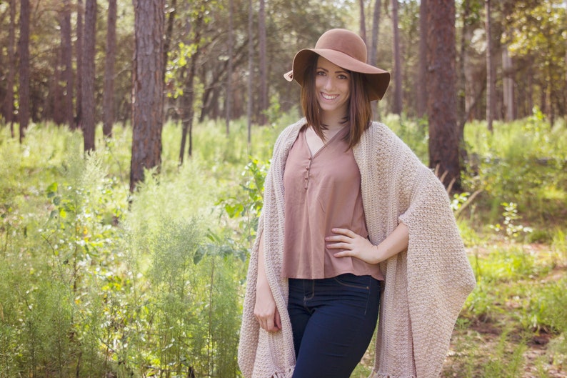 A woman wearing a cream crochet shawl wrap and hat, in a field of greenery.