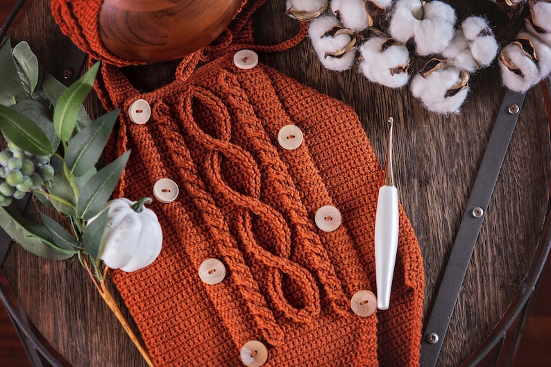 May include: A close-up of a crocheted baby outfit in a rust orange color with wooden buttons. The outfit is lying on a wooden surface with a white crochet hook and a sprig of greenery.