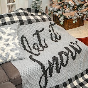 May include: A grey and black crocheted throw blanket with the words "Let it snow" in black lettering. The blanket is on a grey sofa with a white and black snowflake patterned cushion.