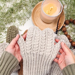 May include: A pair of beige crocheted mittens with a cable knit pattern. The mittens are held by hands wearing green sweaters.