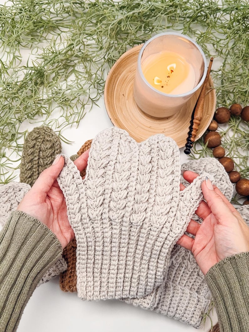 May include: A pair of beige crocheted mittens with a cable knit pattern. The mittens are held by hands wearing green sweaters.