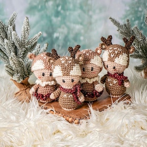 May include: Four crocheted reindeer ornaments in brown and white with red accents. The reindeer are standing on a wooden surface with a fuzzy white background.
